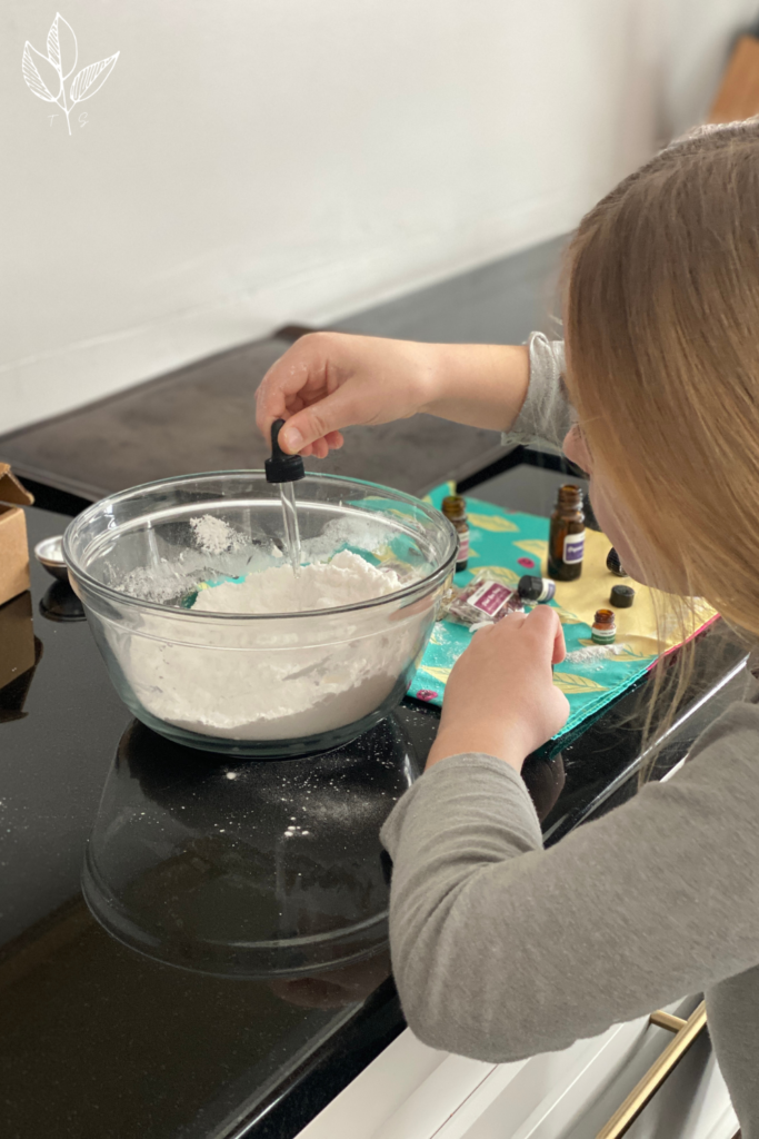 young girl is using a medicine dropper to add essential oil into a large glass mixing bowl filled with the dry ingredients for making bath bombs 