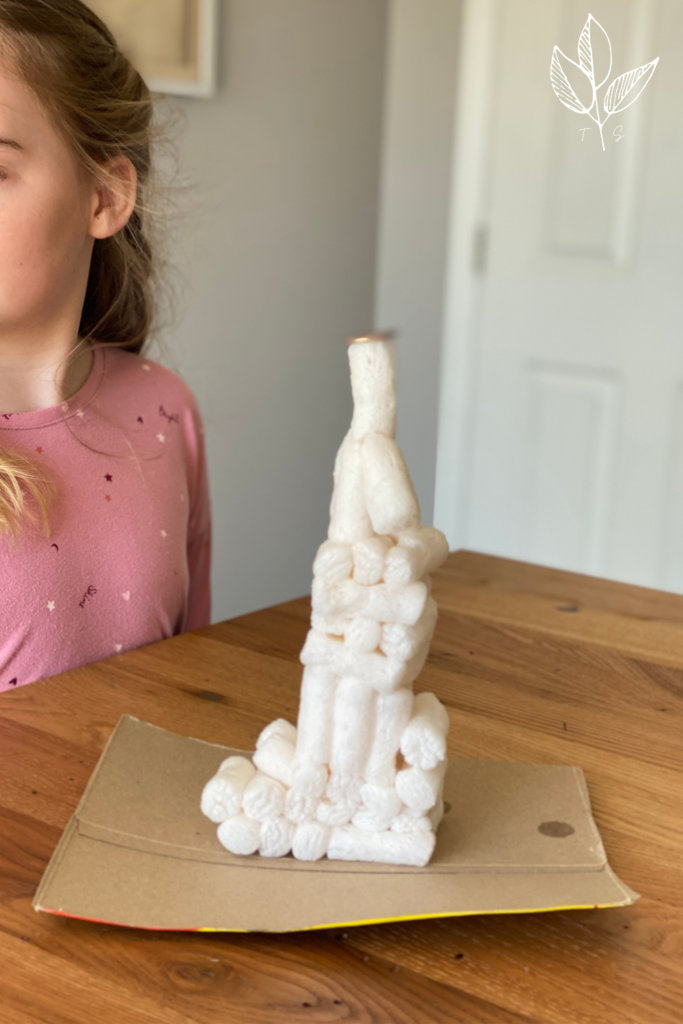 a tower made of compostable packing peanuts sits on top of a piece of cardboard, with a quarter balancing on top of the tower