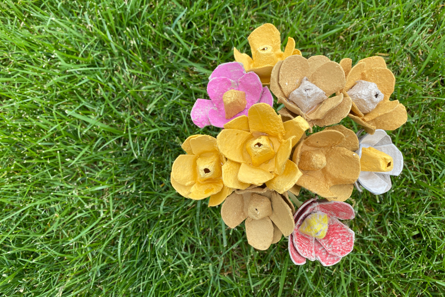 bouquet of yellow, pink and grey egg carton flowers sits in a vase in the green grass.