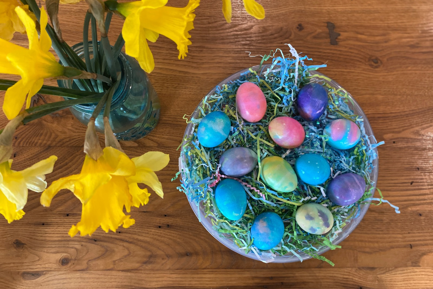 Colorful Dyed eggs sit on a round plate next to daffodils